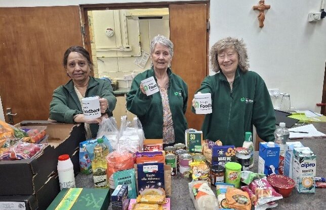 Three volunteers stand with mugs of tea by a table of food items