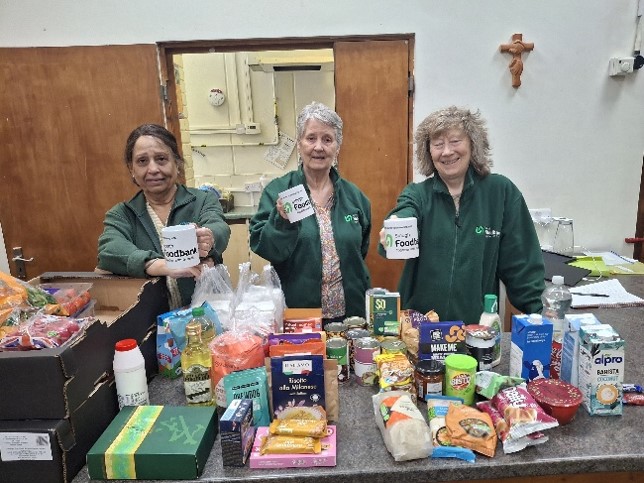 Three volunteers stand with mugs of tea by a table of food items